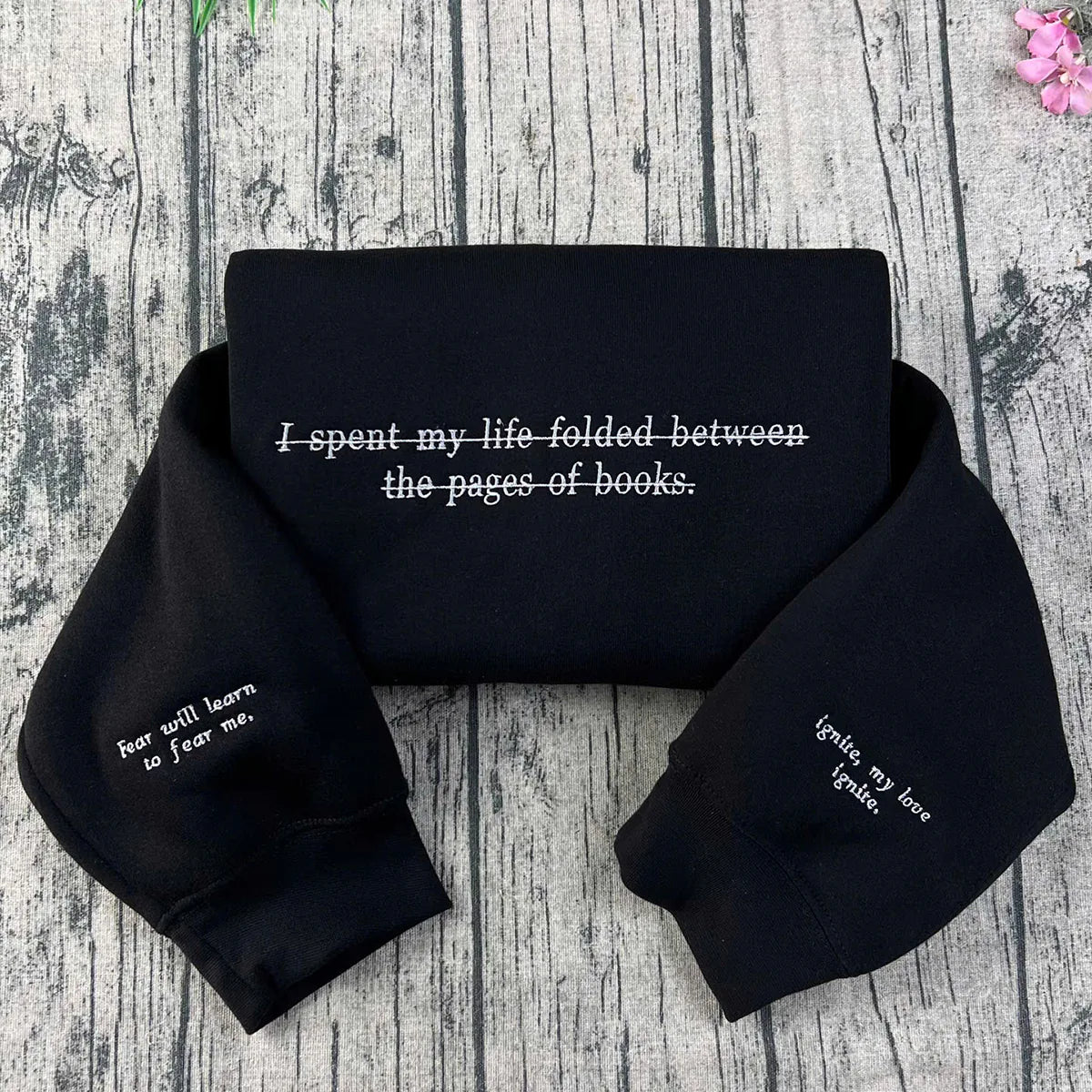 Black crewneck sweatshirt with white embroidered text and quotes from the 'Shatter Me' series, laid flat on a wooden background with a pink flower at the top right corner.