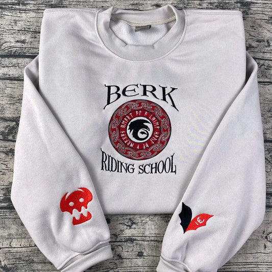 White sweatshirt with 'Berk Riding School' design on a wooden surface