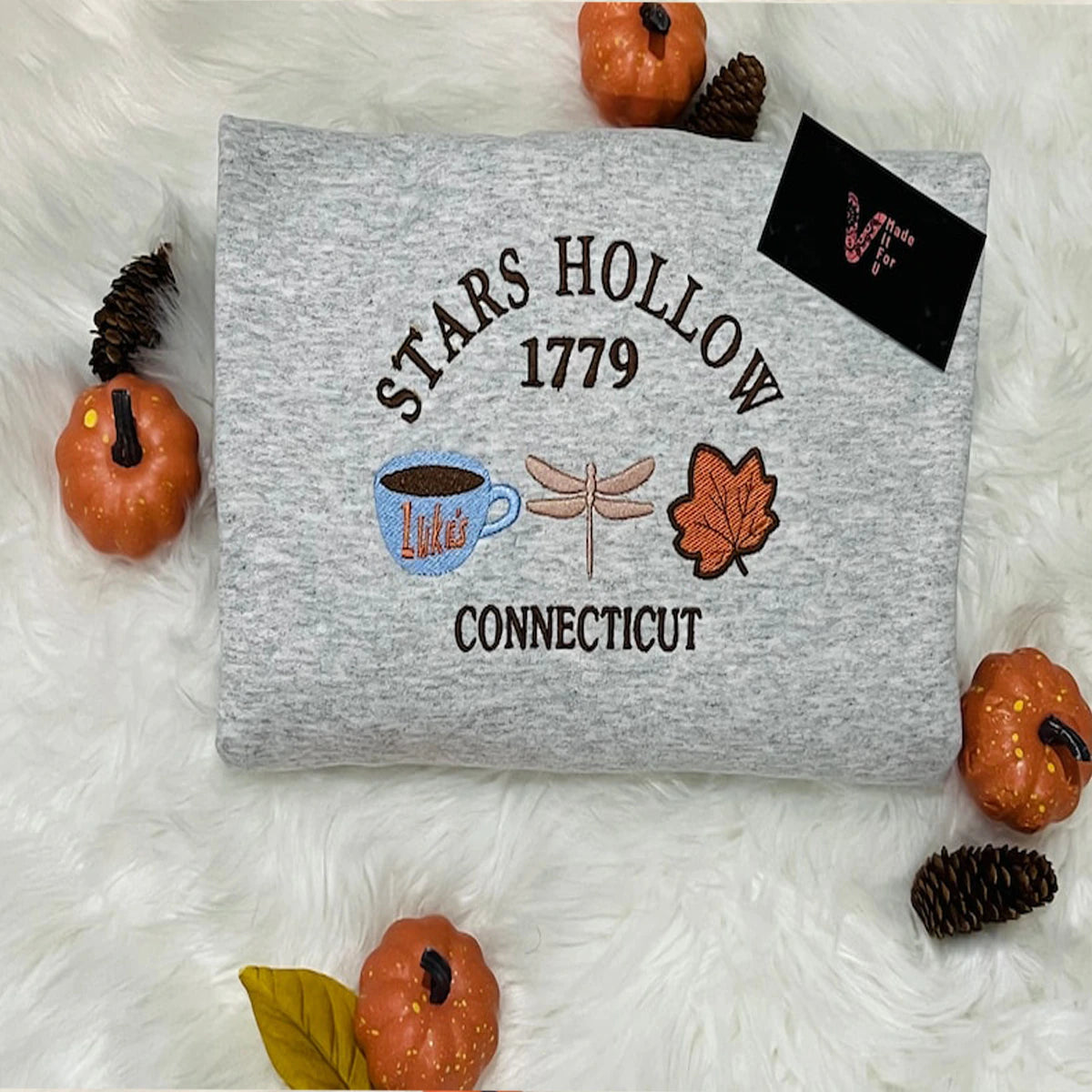 Gray sweatshirt with 'Stars Hollow 1779 Connecticut' design on a white surface with decorative pumpkins and pinecones.