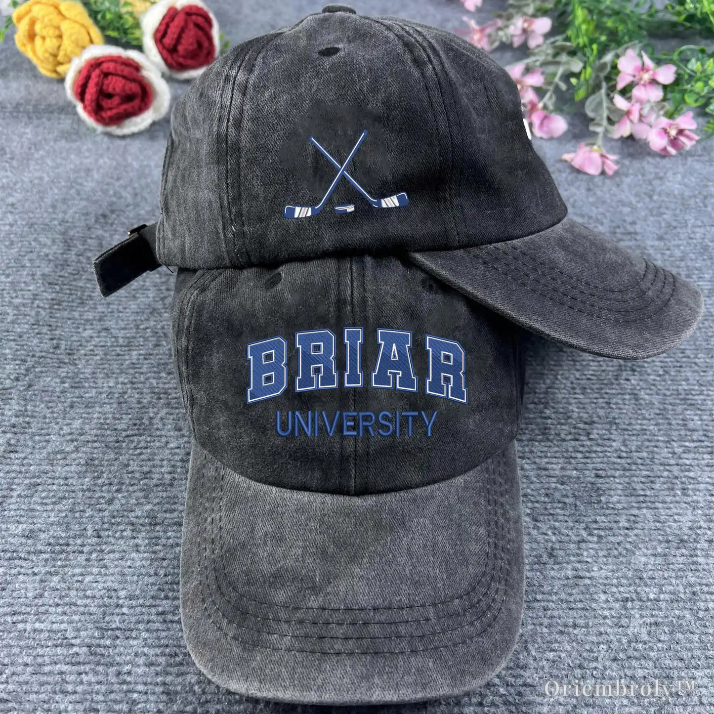 Black baseball cap with 'BRIAR UNIVERSITY' text on a textured surface with flowers in the background