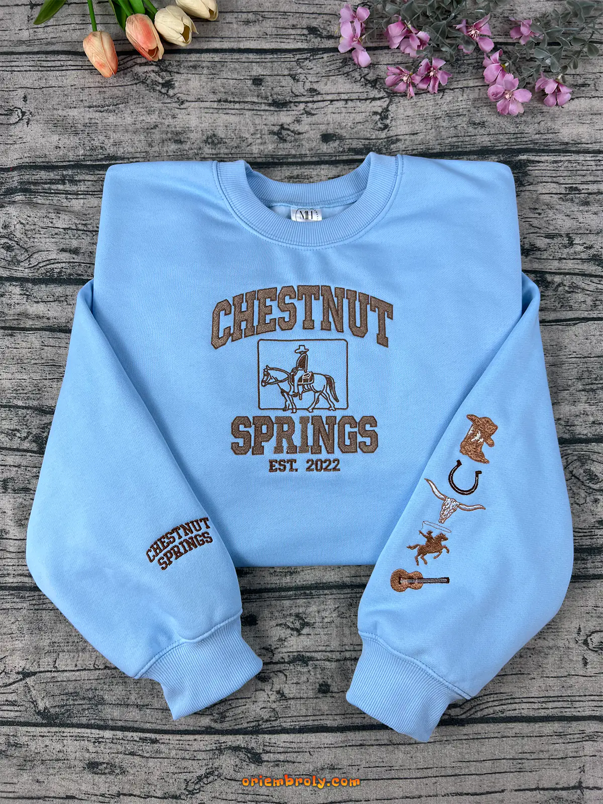 Light blue sweatshirt with 'Chestnut Springs' design on a wooden surface with flowers.