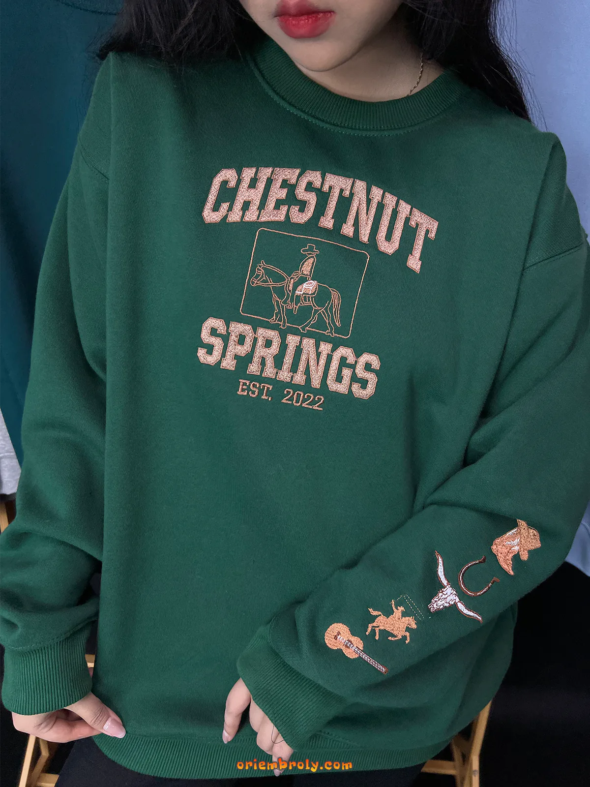 Green sweatshirt with 'Chestnut Springs' text and embroidery on a person against a blue wall.