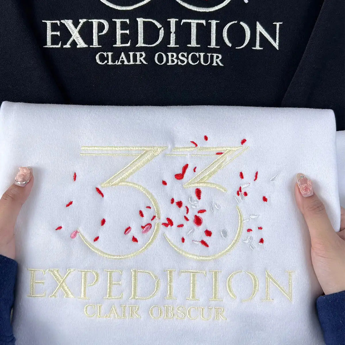Embroidered design with '33 Expedition Clair Obscur' on a white fabric held by hands.