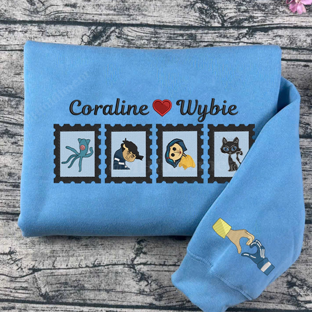 Folded blue sweatshirt with character illustrations and 'Coraline Wybie' text on a wooden surface