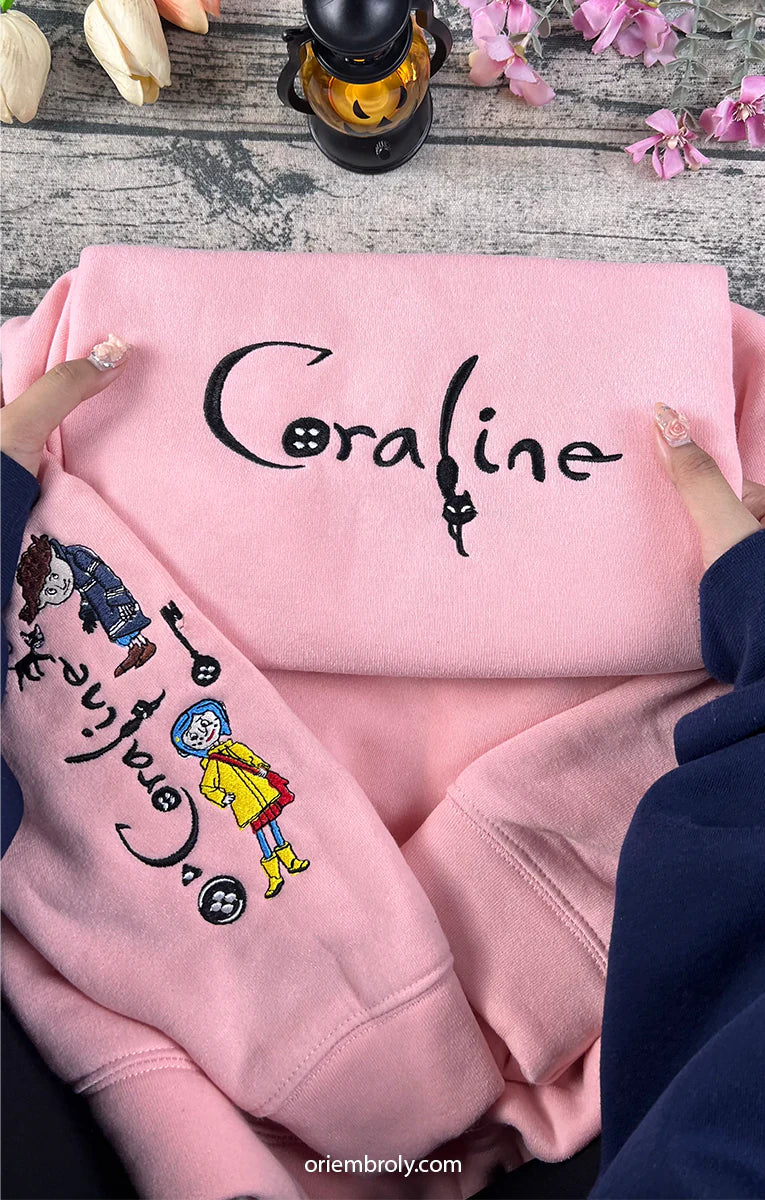 Pink hoodie with 'Coraline' design held by hands on a wooden surface with flowers.