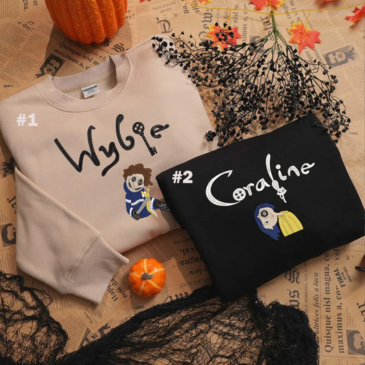 Two sweatshirts with embroidered names and characters on a textured surface with pumpkins and flowers.