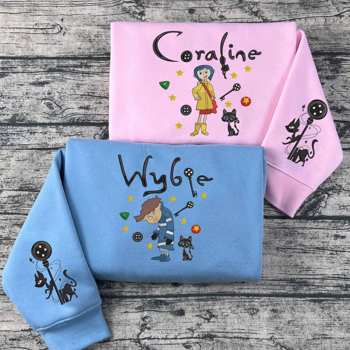 Two hoodies, one pink and one blue, with embroidered characters and text on a wooden surface.