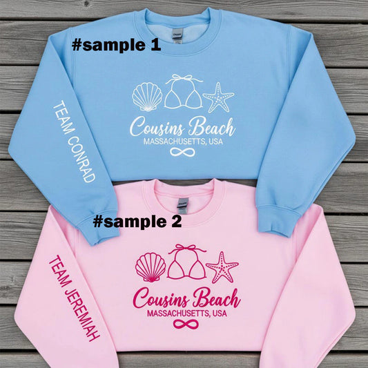 Two sweatshirts, one blue and one pink, with 'Cousins Beach' text and graphics on a wooden surface.