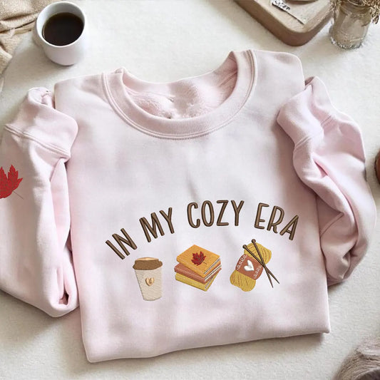 Pink sweatshirt with 'In My Cozy Era' text and illustrations on a light surface.