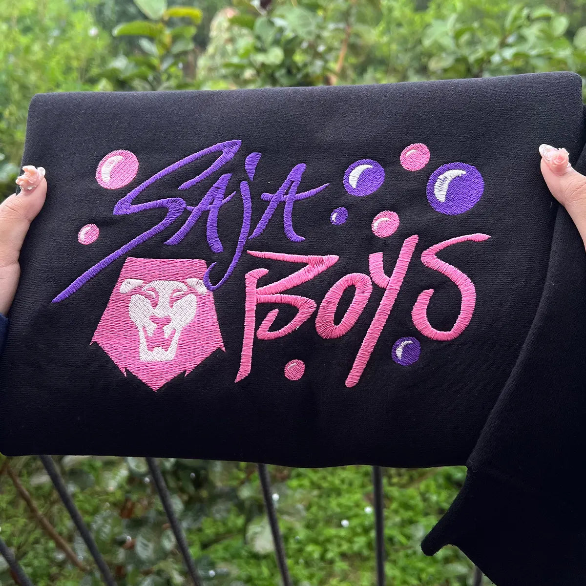 Black sweatshirt with 'Saja Boys' text and logo held by a person outdoors.