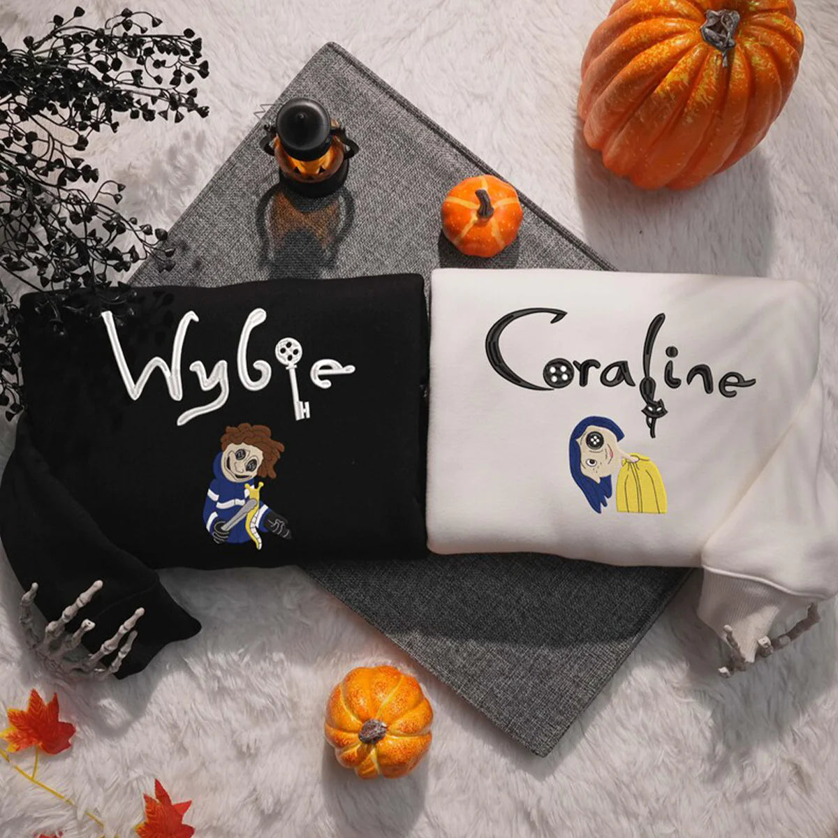 Two hoodies with 'Wybie' and 'Coraline' designs on a textured surface with pumpkins and leaves.