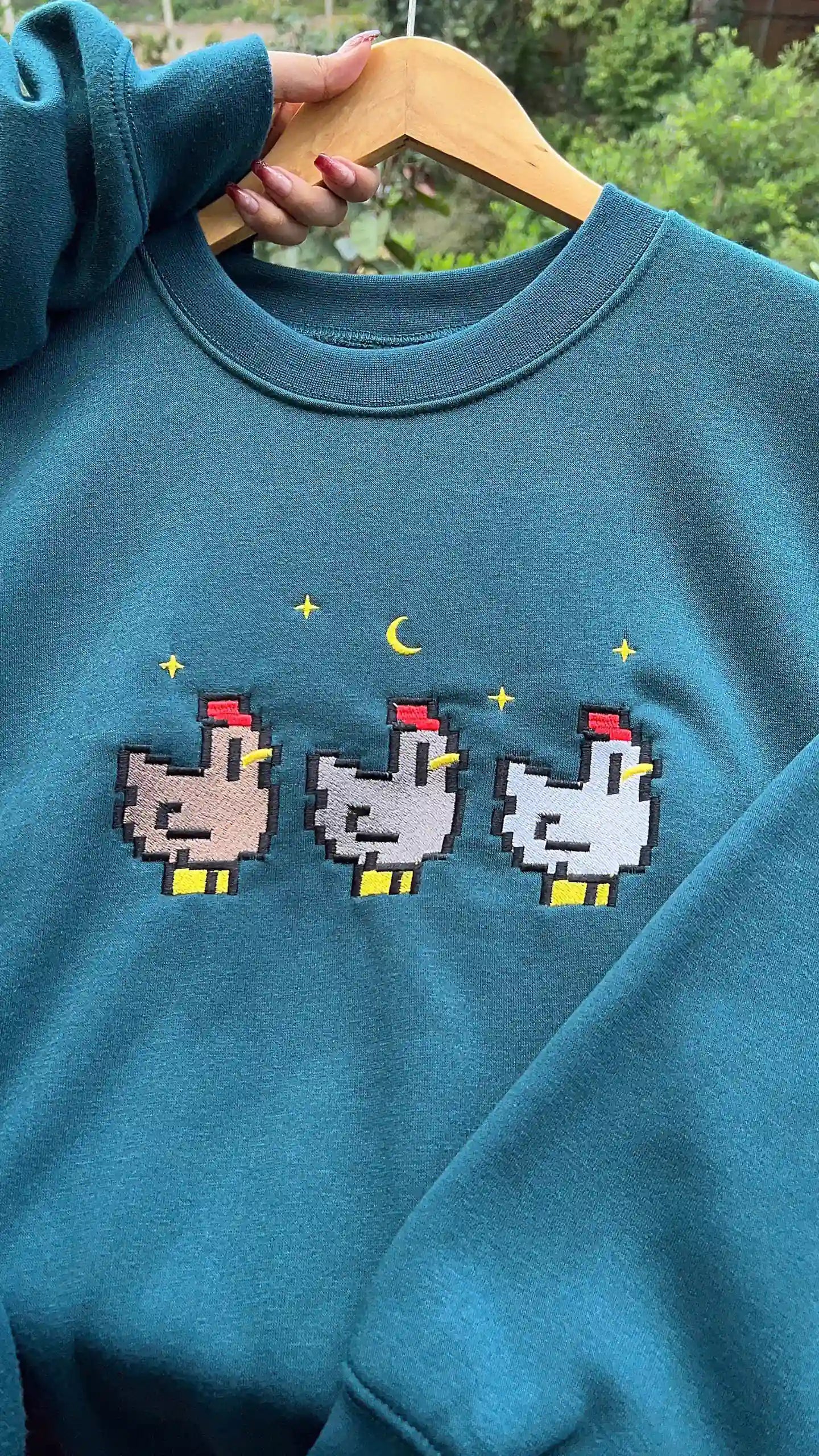Teal sweatshirt with pixelated chicken design on a hanger
