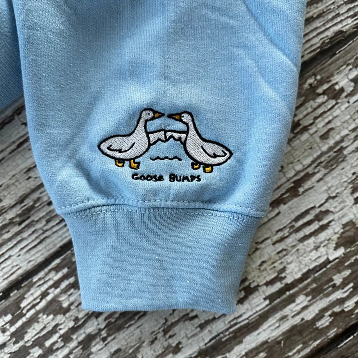 Light blue sweatshirt sleeve with embroidered design of two ducks and 'GOOSE BUMPS' text on a wooden surface.