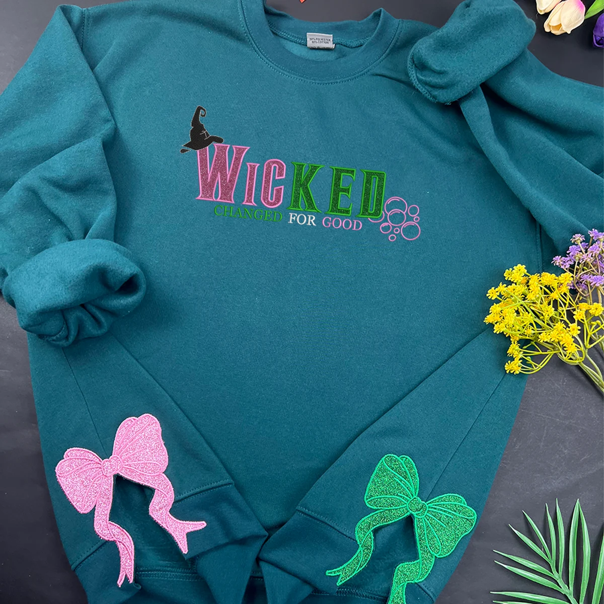Teal sweatshirt with 'WICKED' text and decorative elements on a dark surface with flowers and leaves.