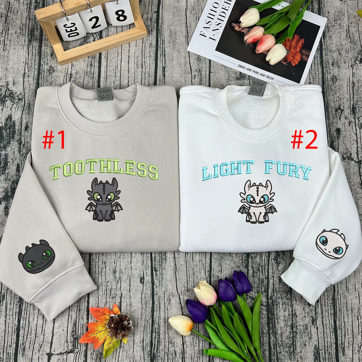 Two folded sweatshirts with embroidered designs on a wooden surface with flowers and a calendar.