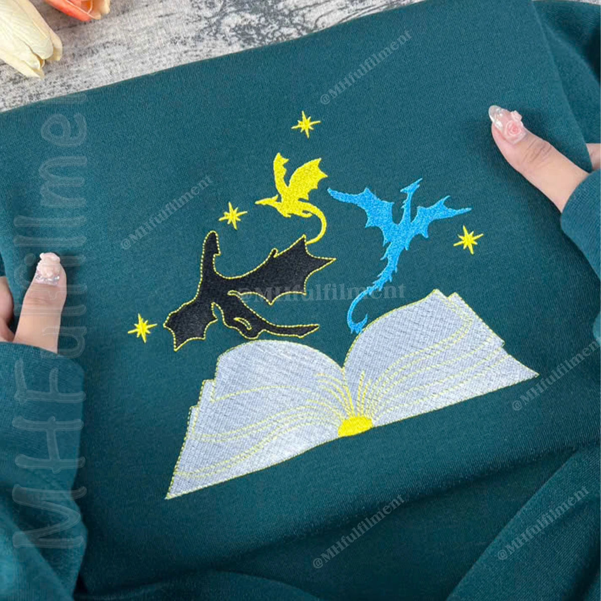Embroidered design of dragons and an open book on a teal fabric.