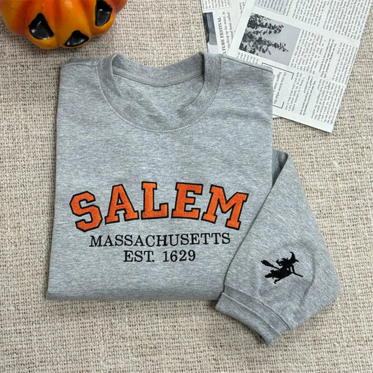 Folded gray sweatshirt with 'Salem Massachusetts Est. 1629' text on a textured surface with a pumpkin and newspaper in the background.