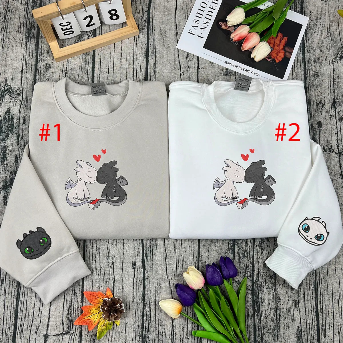 Two folded sweatshirts with dragon designs on a wooden surface with flowers and a calendar.