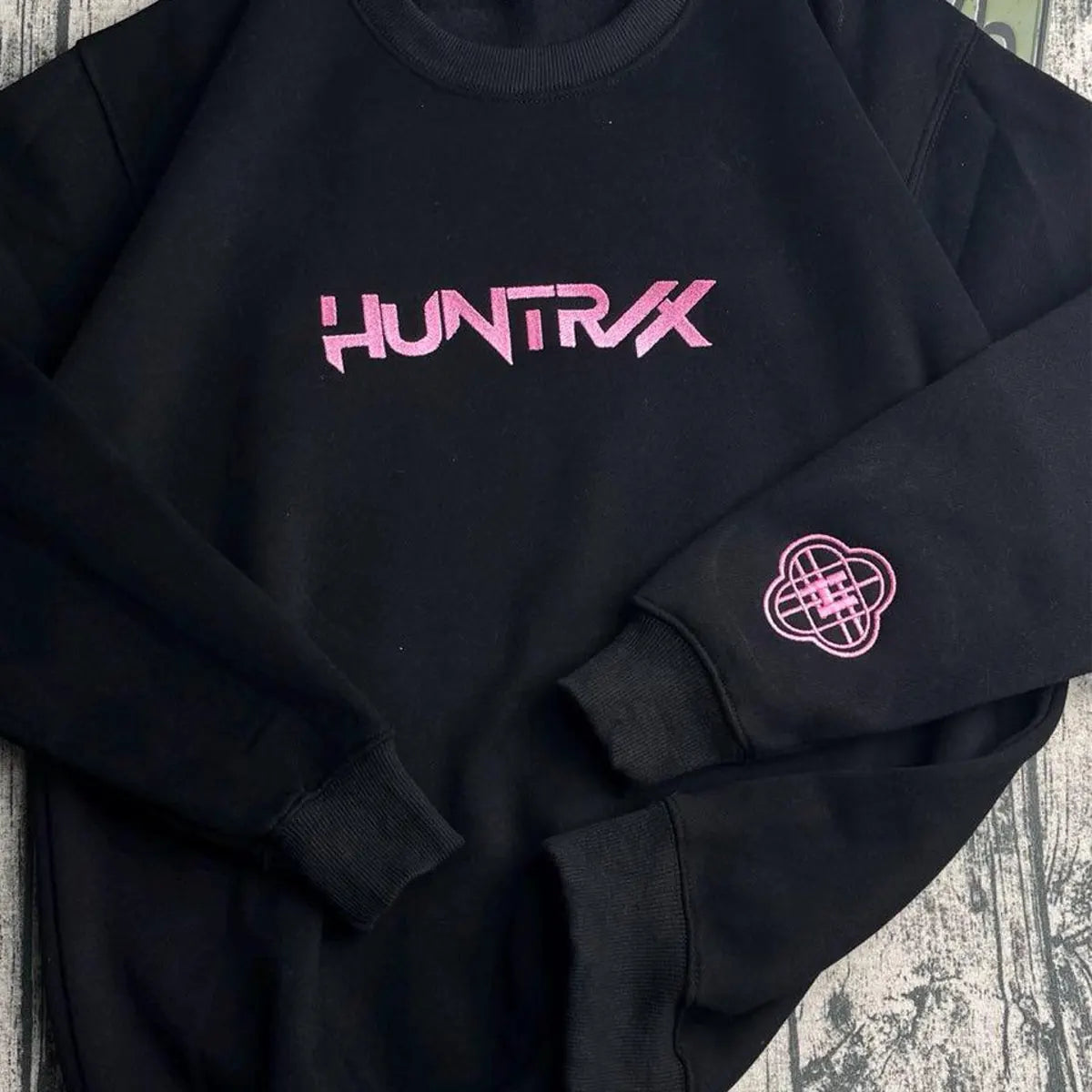 Black sweatshirt with pink 'HUNTRX' logo on a textured surface