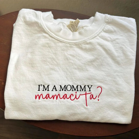 Folded white sweatshirt with text 'I'm a mommy mamacita?' on a wooden surface