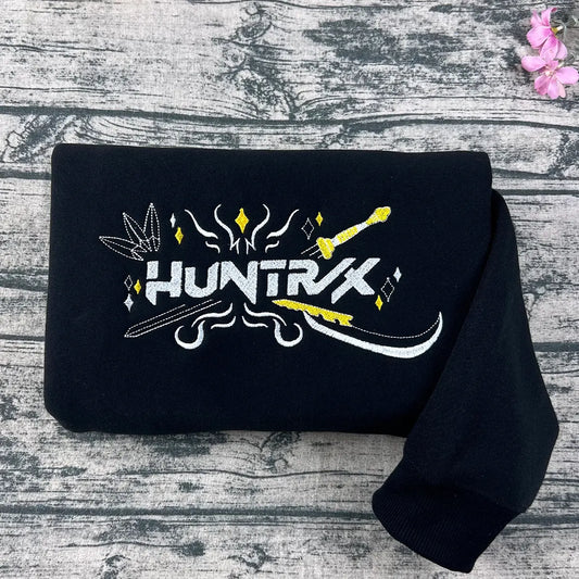 Black t-shirt with 'HUNTRX' logo on a wooden surface with flowers.