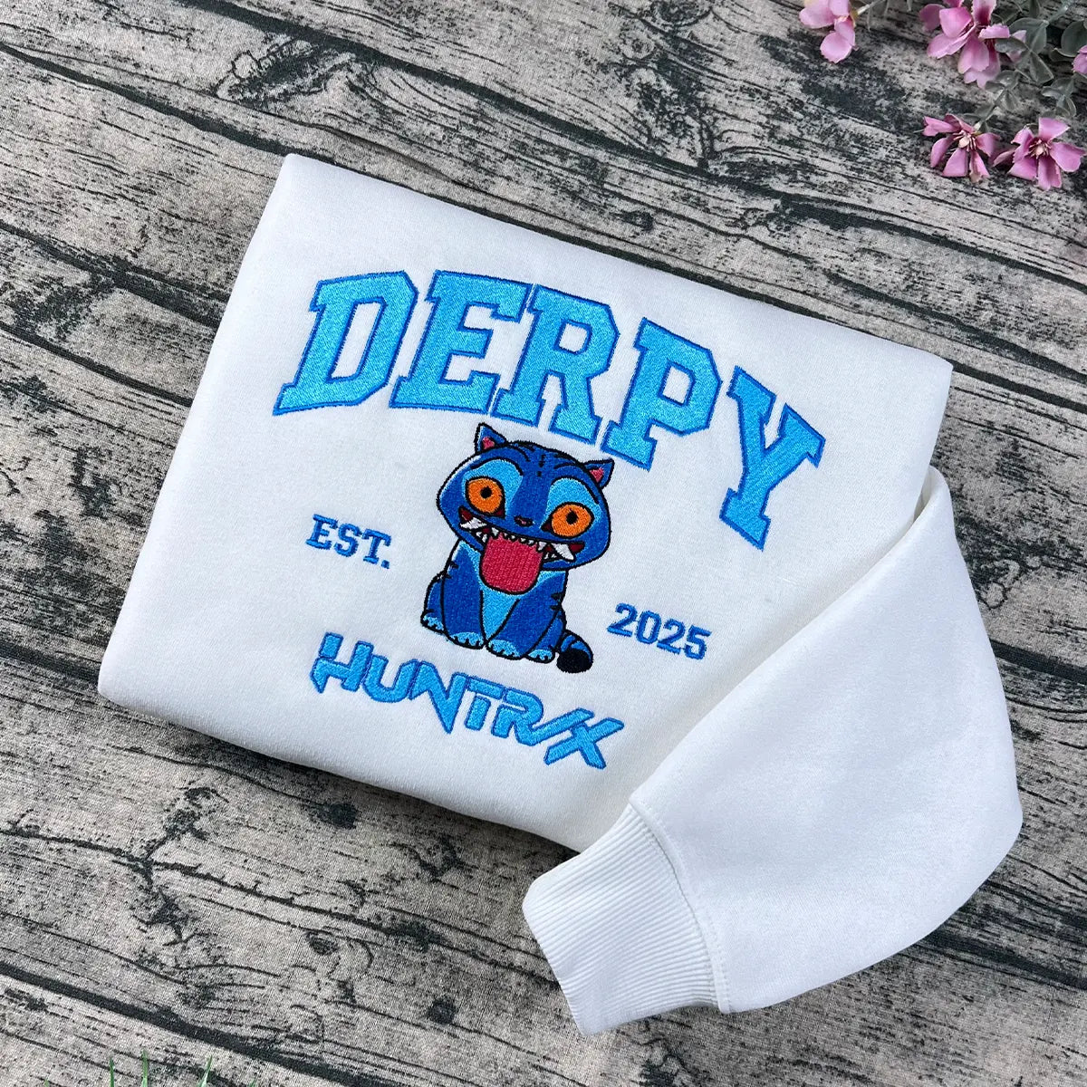 White sweatshirt with 'DERPY' and cartoon character on a wooden surface with flowers.