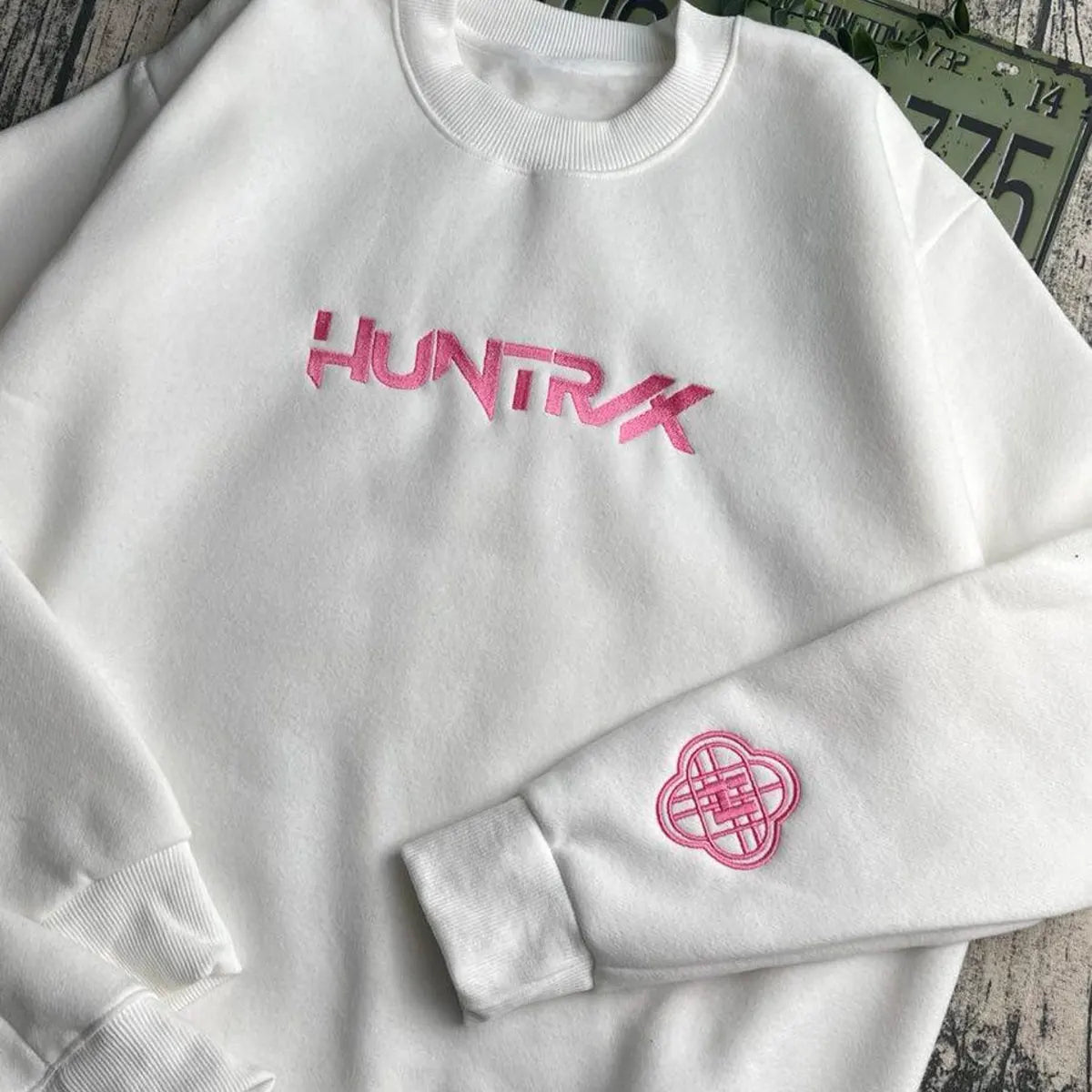 White sweatshirt with pink 'HUNTRX' logo on a wooden surface