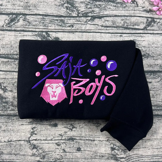 Black sweatshirt with 'SAJA Boys' text and logo on a wooden surface