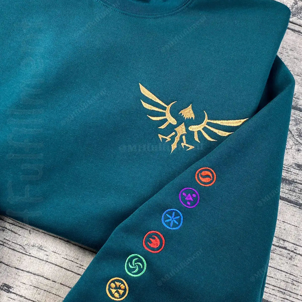 Teal hoodie with gold emblem and colorful symbols on a wooden surface