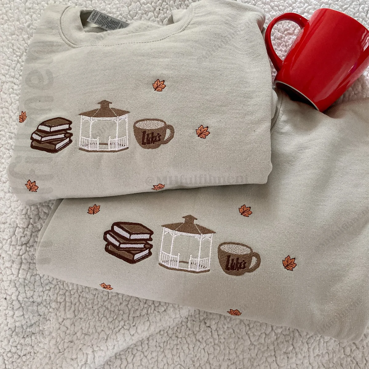 Folded light gray sweatshirt with embroidered designs on a textured surface with a red mug.