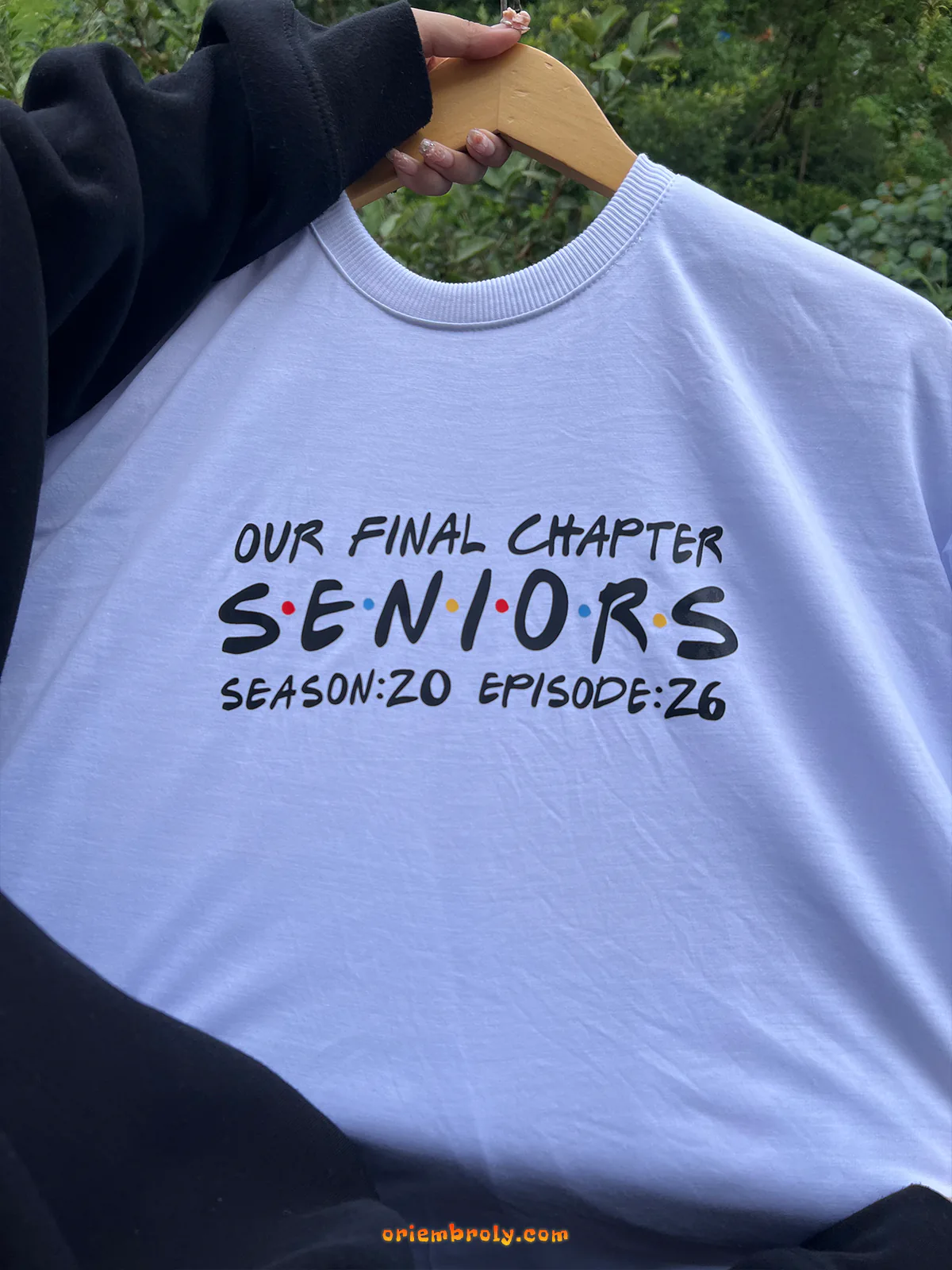 White sweatshirt with 'Our Final Chapter Seniors' text on a hanger against a blurred green background