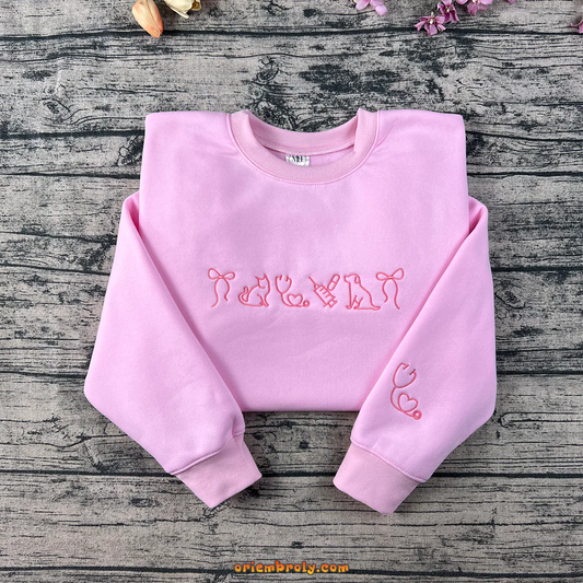 Pink embroidered sweatshirt featuring cat, dog, syringe, stethoscope, and heart designs β perfect for veterinarians or pet lovers.