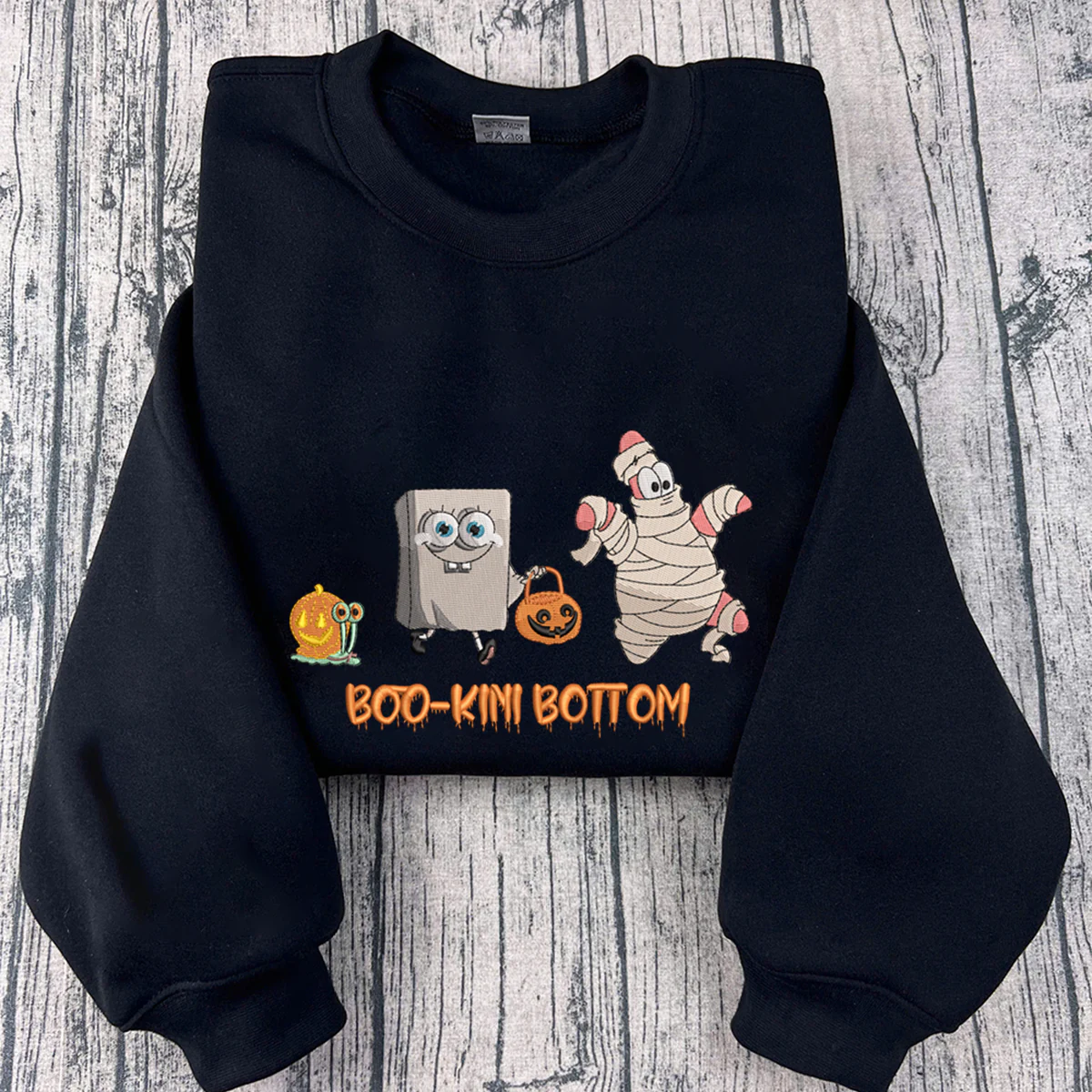 Black sweatshirt with cartoon characters and 'BOO-KINI BOTTOM' text on a wooden surface