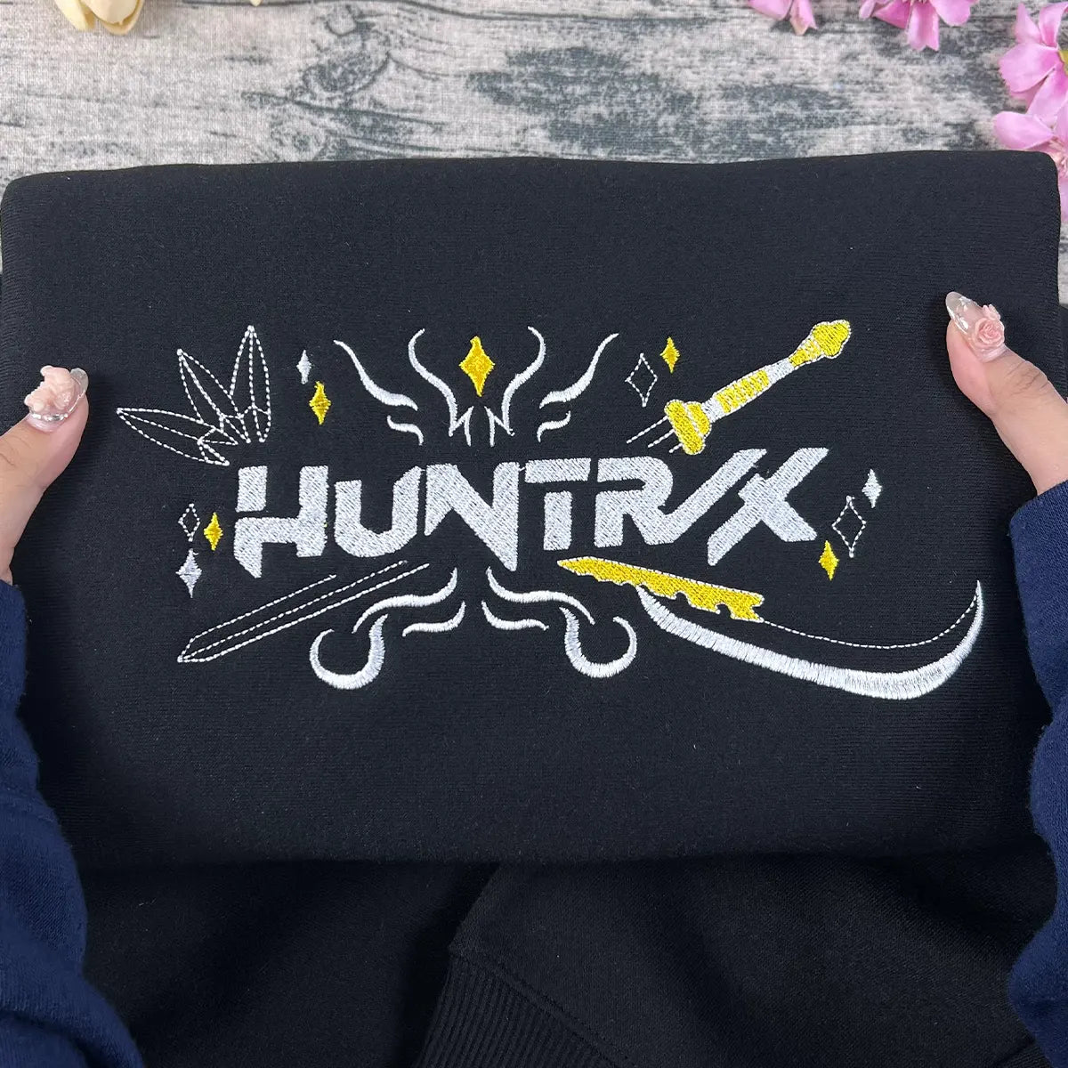Black fabric with embroidered design and 'HUNTRK' text held by hands on a wooden surface.