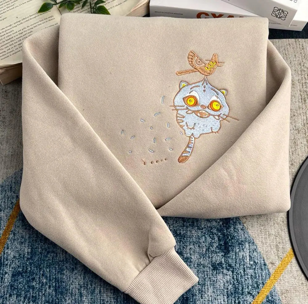 Beige sweatshirt with embroidered cat design on a textured surface
