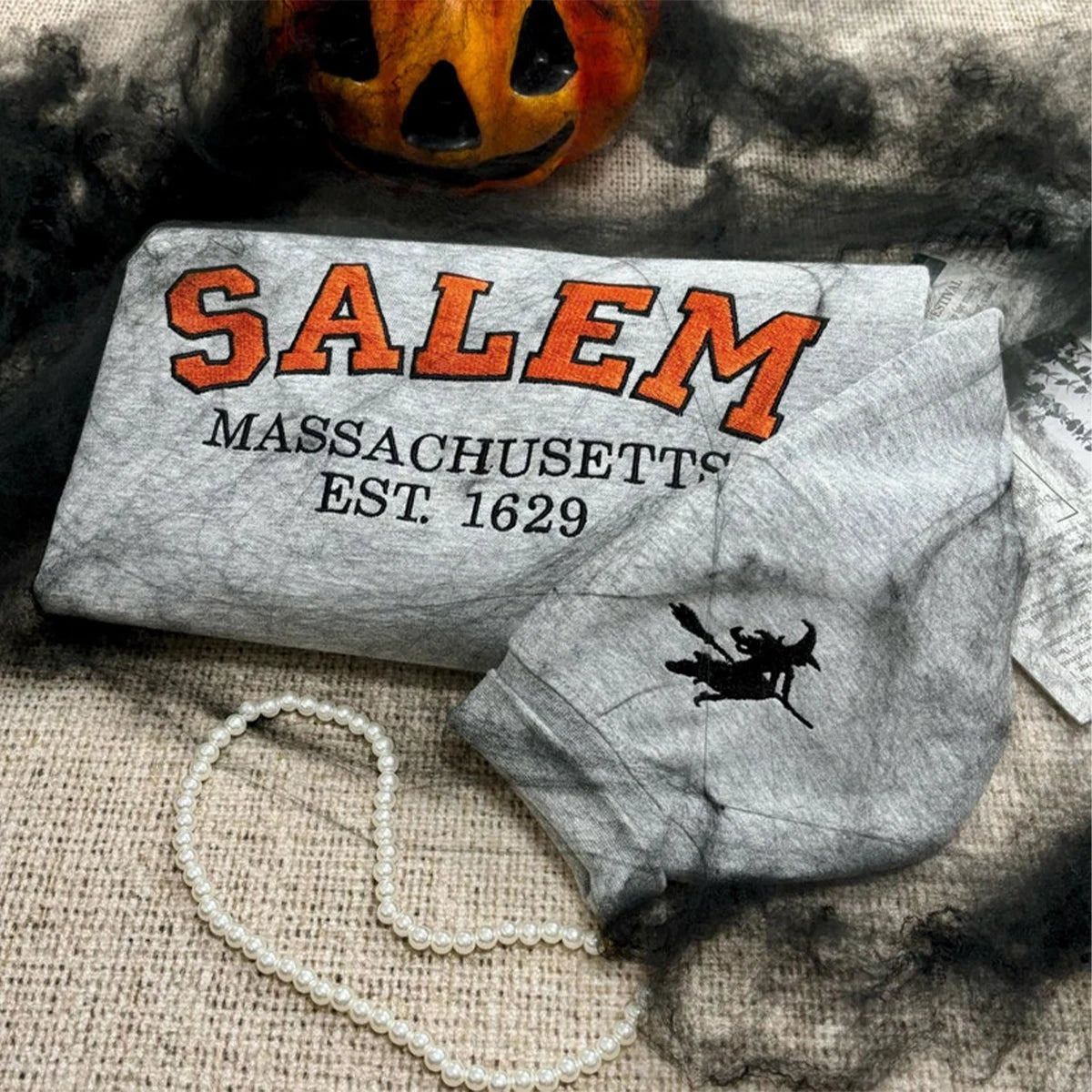 Gray face mask with 'Salem Massachusetts Est. 1629' text and witch design, Halloween pumpkin in background.