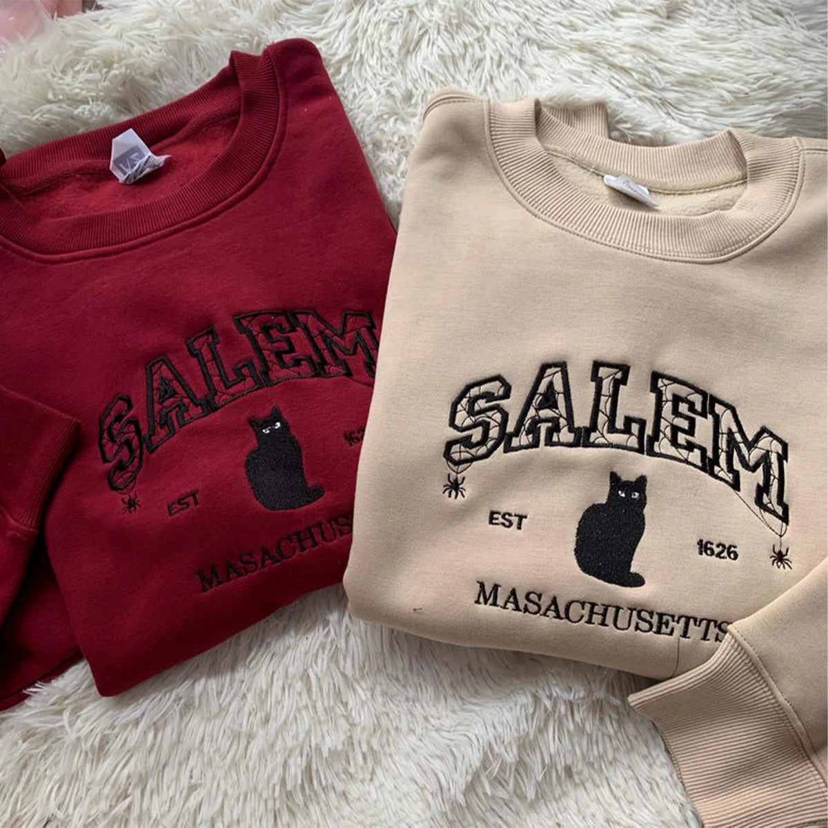 Two sweatshirts with 'Salem, Massachusetts' text and a black cat graphic on a fluffy white surface.