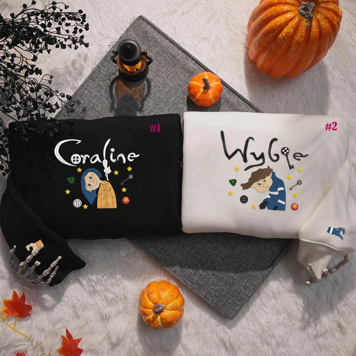 Two hoodies with embroidered designs and names on a textured surface with pumpkins and leaves.