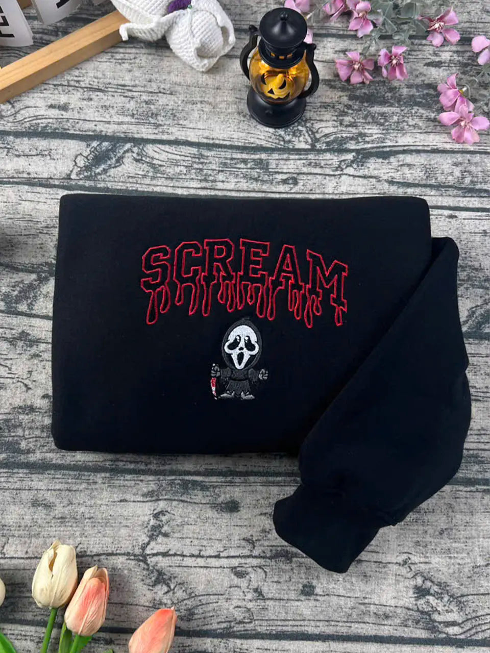 Black hoodie with 'Scream' text and logo on a wooden surface with flowers and a lantern.