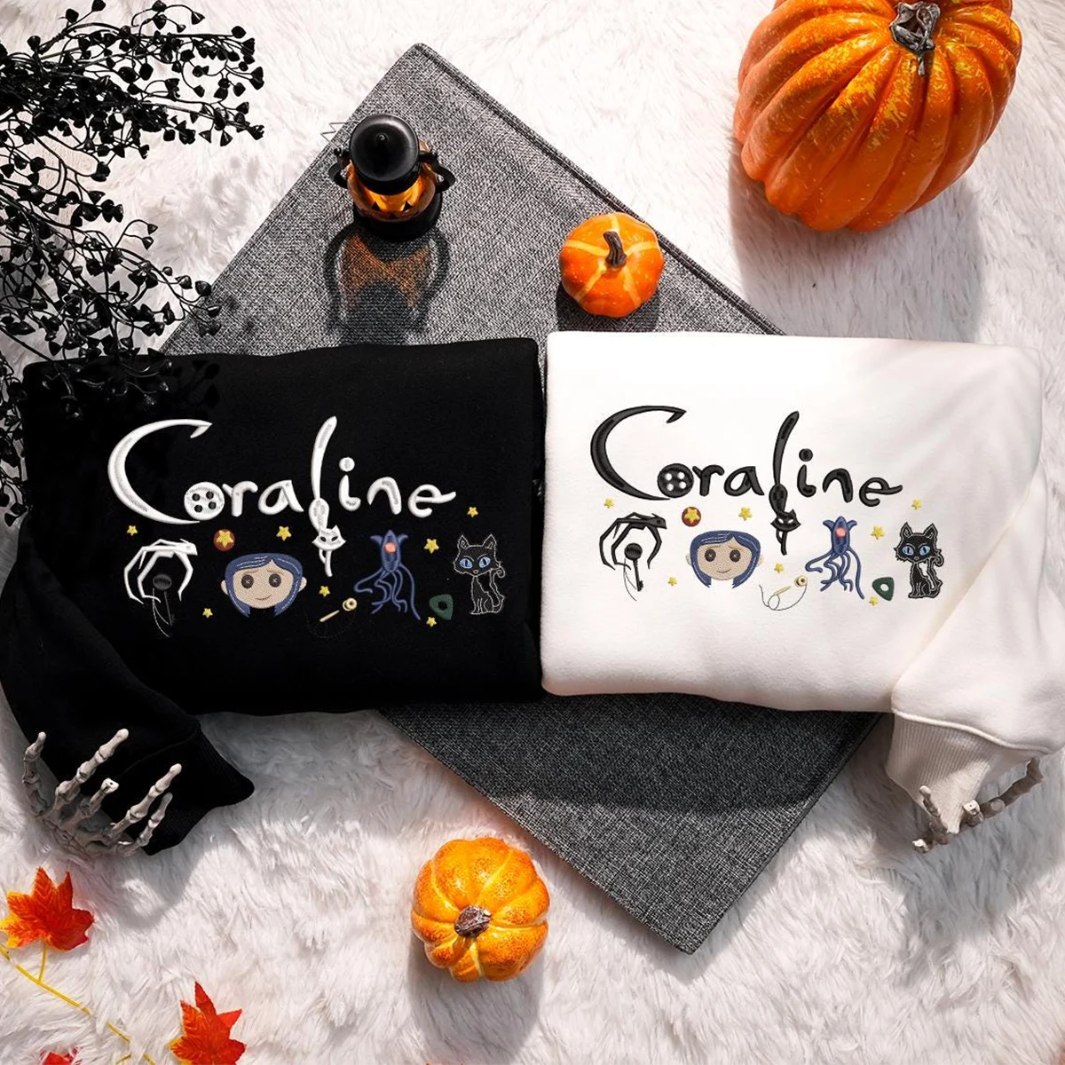 Black and white sweatshirts with 'Coraline' design on a textured surface with pumpkins and leaves.