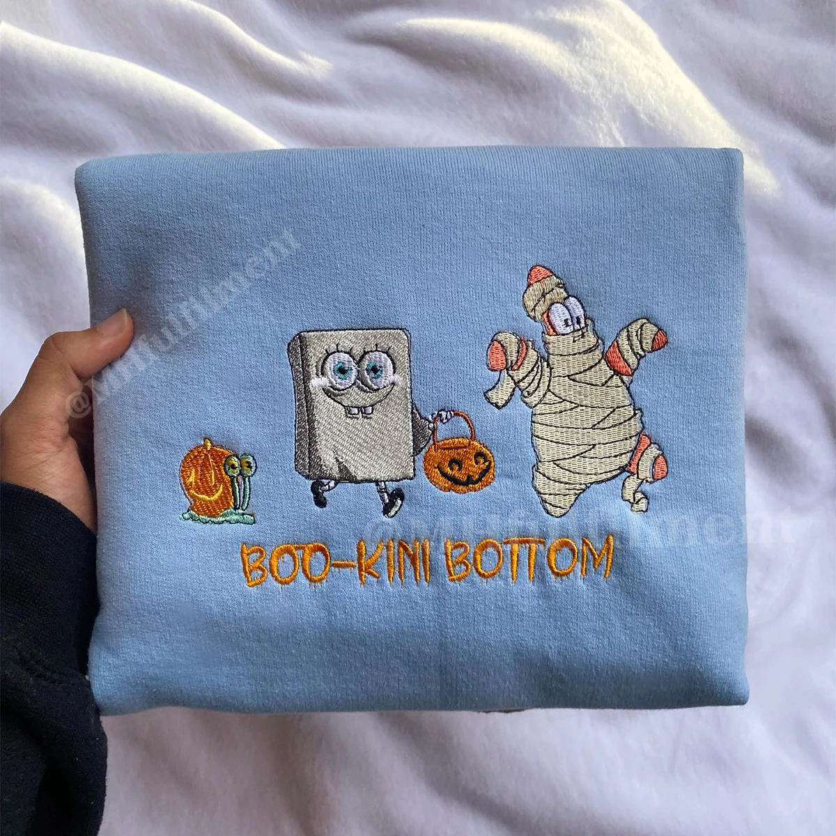 Embroidered fabric with cartoon characters and 'BOO-KINI BOTTOM' text on a light blue background.