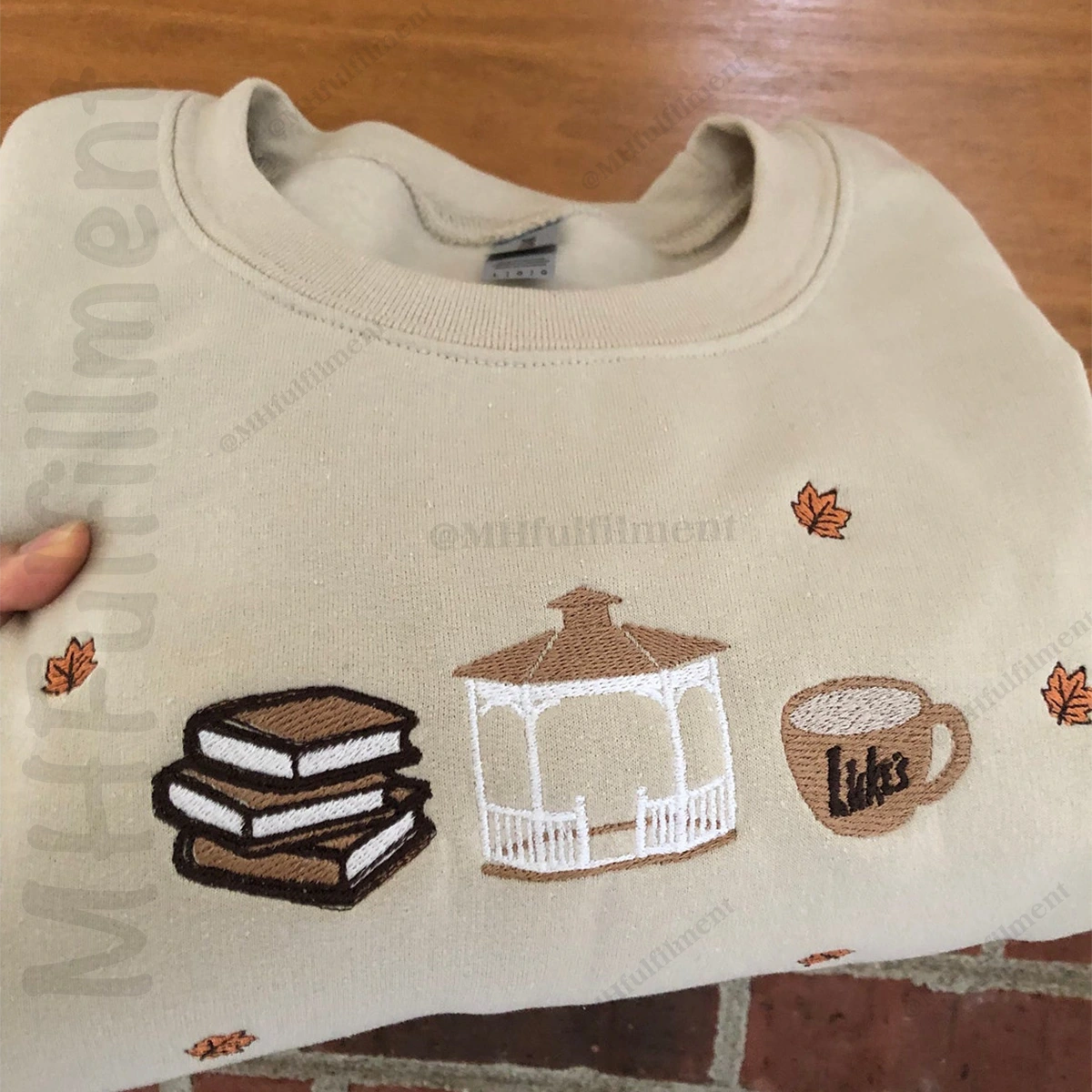 Beige sweatshirt with embroidered designs of books, a gazebo, and a coffee cup on a wooden surface.