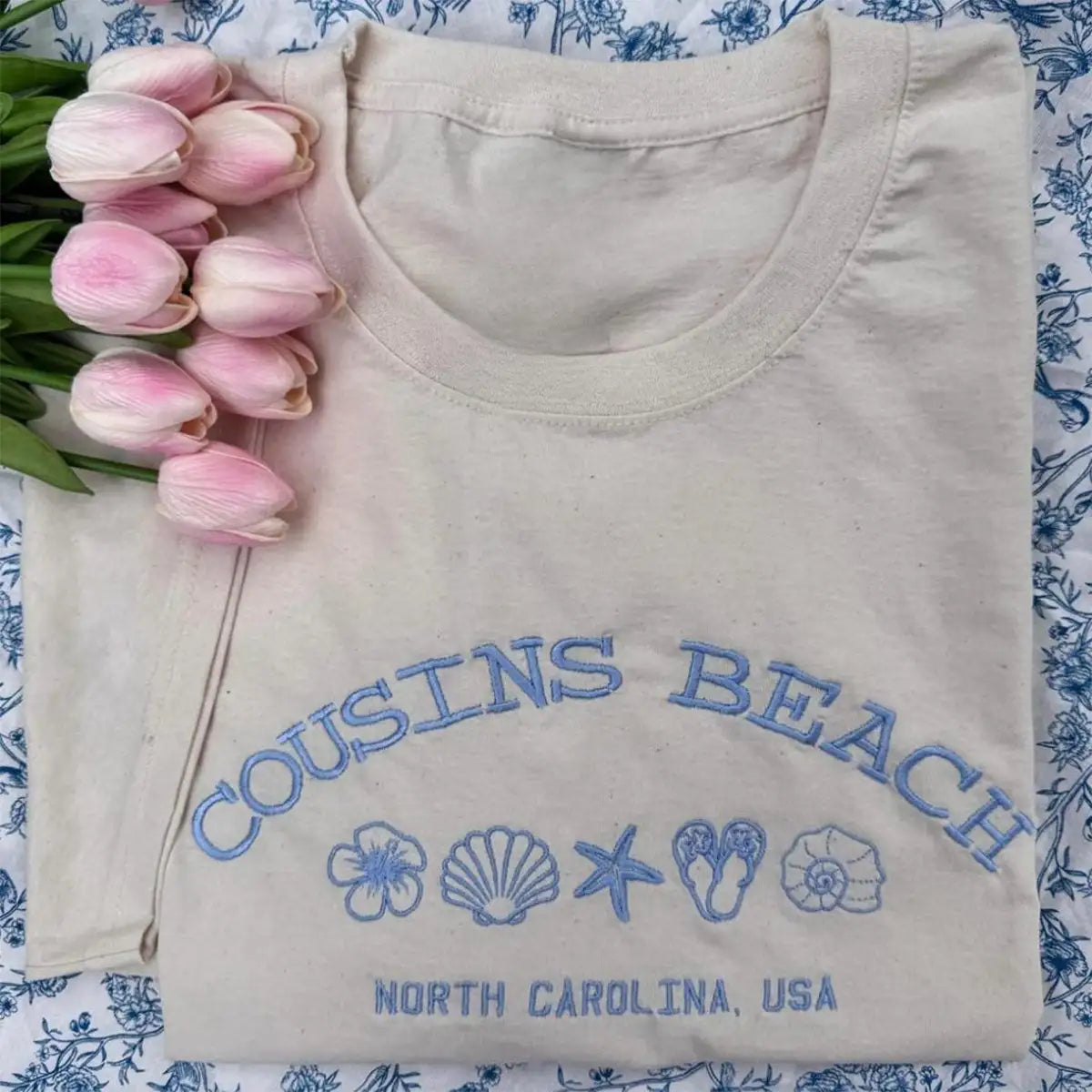 Folded beige shirt with 'Cousins Beach, North Carolina, USA' text and graphics on a floral background.