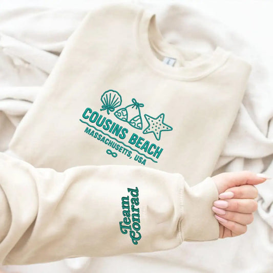 Beige sweatshirt with teal 'Cousins Beach' and 'Team Conrad' text on a white background