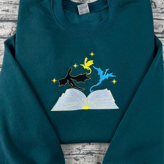 Teal hoodie with embroidered design of a book, cat, and bird on a wooden surface