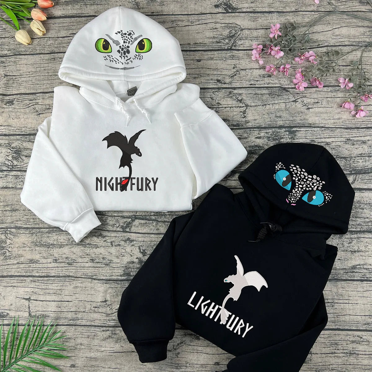 Two hoodies with dragon designs and text on a wooden surface with flowers.