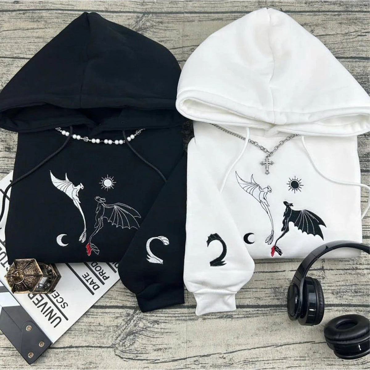 Black and white hoodies with dragon designs on a wooden surface with headphones and a magazine.