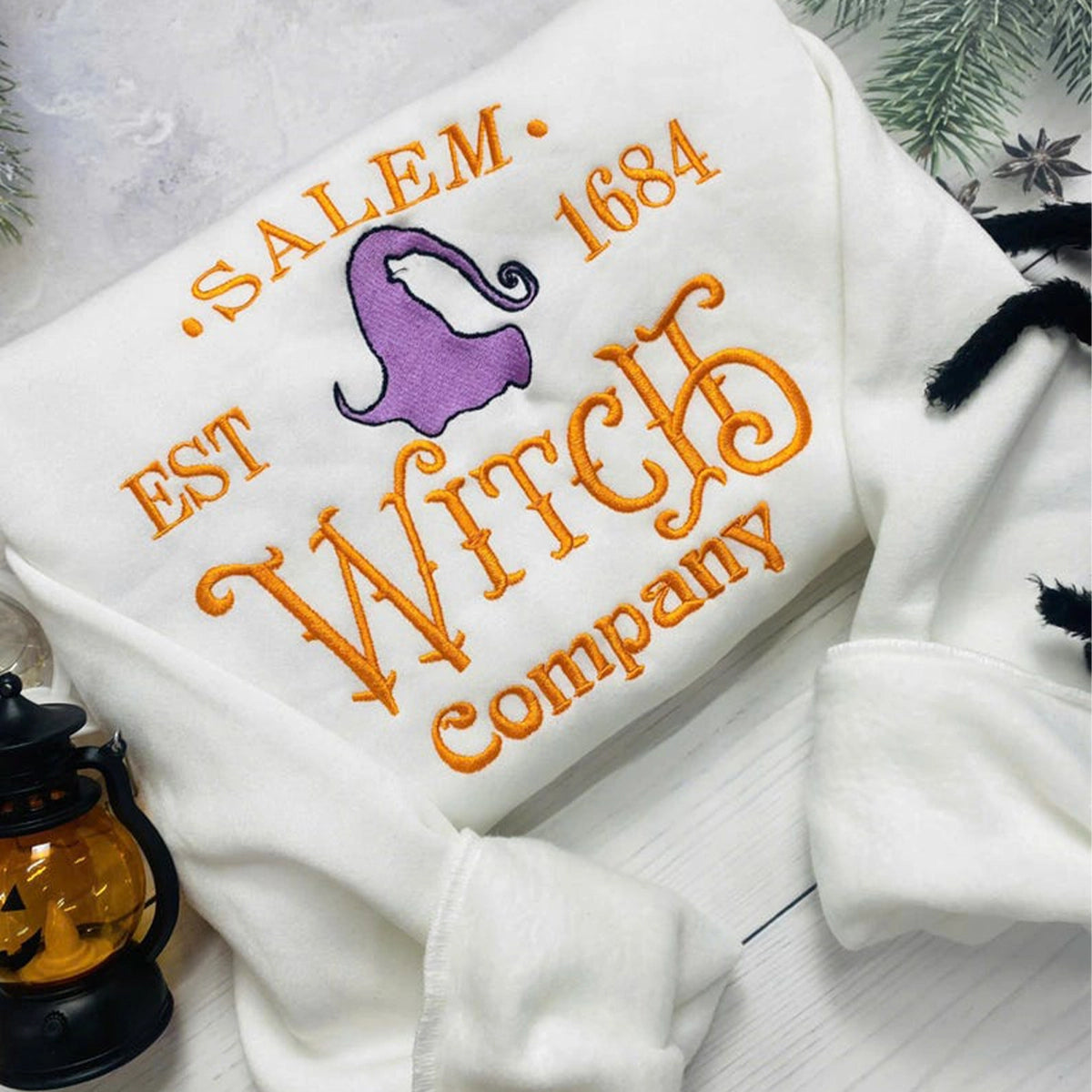 White sweatshirt with embroidered text and witch hat design on a light background.