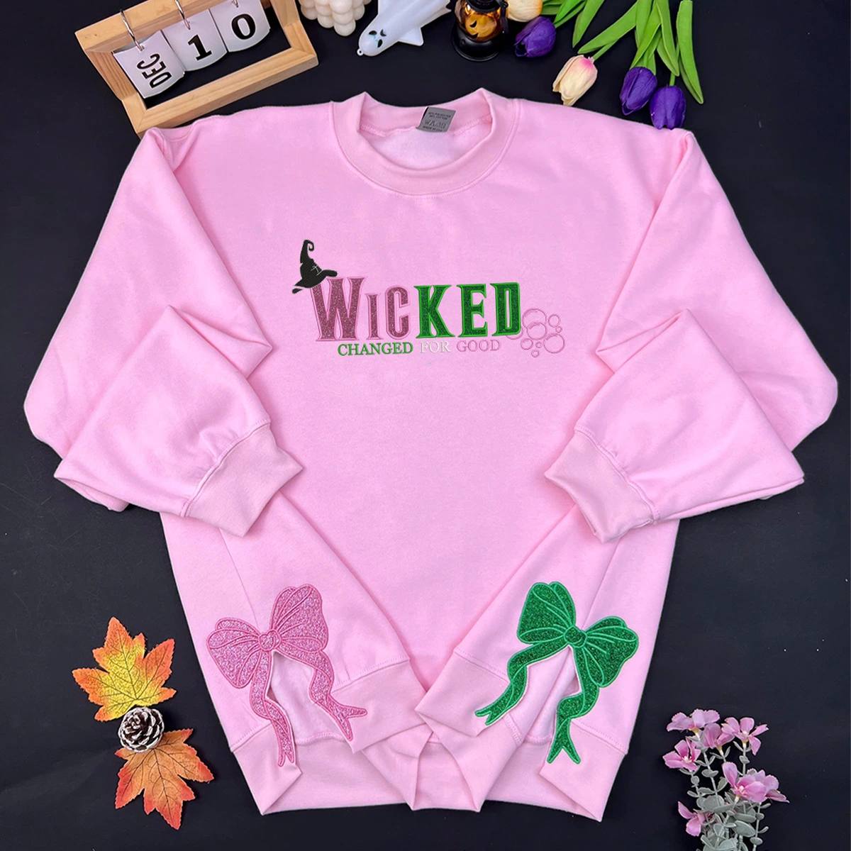 Pink sweatshirt with 'WICKED' text and decorative elements on a dark surface with flowers and a calendar.