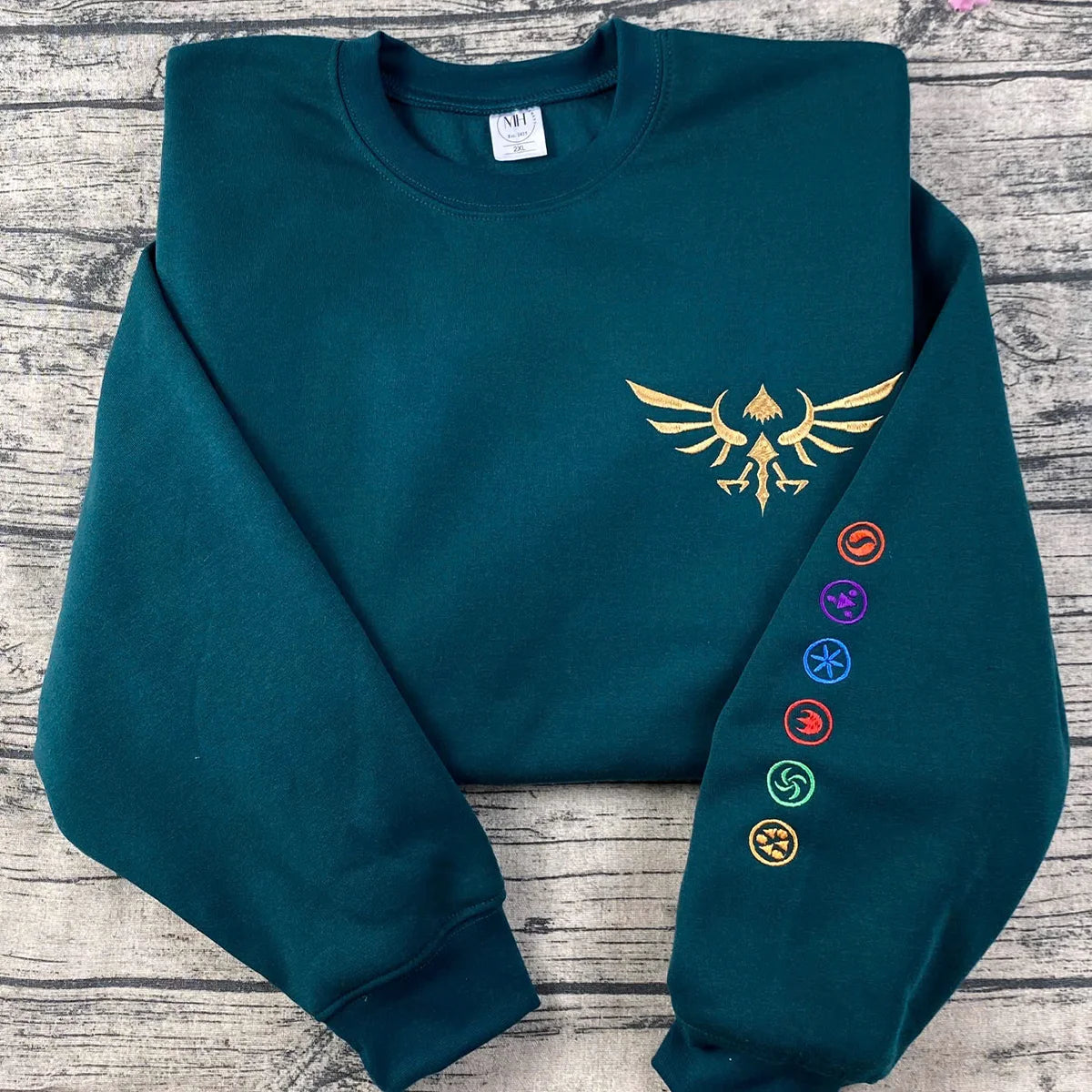 Teal sweatshirt with gold emblem and colorful symbols on a wooden surface